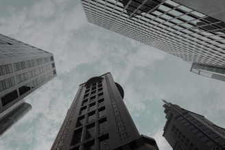 Skyscrapers rise toward a cloudy sky from an upward perspective, displaying modern architectural designs and glass facades.