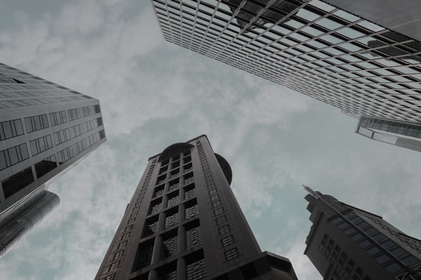 Skyscrapers rise toward a cloudy sky from an upward perspective, displaying modern architectural designs and glass facades.