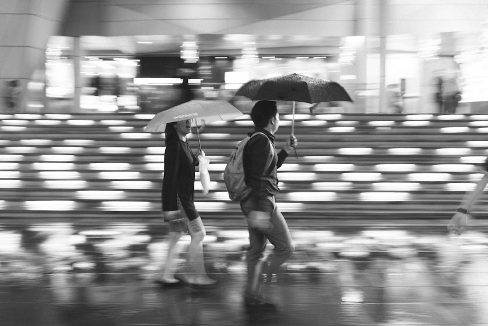 grayscale photography of man and woman walking while holding umbrellas