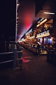 A nighttime scene featuring a row of brightly lit restaurants along a waterfront walkway. The signs of the restaurants are illuminated, casting a warm glow. Tables and chairs are arranged outside the establishments, and a person is seen near the entrance of one of the restaurants. The walkway is bordered by a metal railing, separating it from the body of water on the left. Neon and street lights contribute to the vibrant atmosphere.