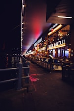 A nighttime scene featuring a row of brightly lit restaurants along a waterfront walkway. The signs of the restaurants are illuminated, casting a warm glow. Tables and chairs are arranged outside the establishments, and a person is seen near the entrance of one of the restaurants. The walkway is bordered by a metal railing, separating it from the body of water on the left. Neon and street lights contribute to the vibrant atmosphere.