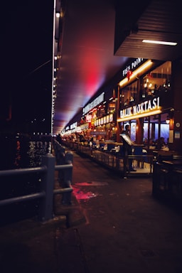 A nighttime scene featuring a row of brightly lit restaurants along a waterfront walkway. The signs of the restaurants are illuminated, casting a warm glow. Tables and chairs are arranged outside the establishments, and a person is seen near the entrance of one of the restaurants. The walkway is bordered by a metal railing, separating it from the body of water on the left. Neon and street lights contribute to the vibrant atmosphere.