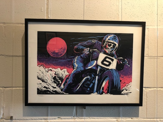 A framed artwork depicts a skeleton in a motorcycle suit riding a motorbike with the number 6 on the front. The scene is dramatic and dynamic, with a large red moon and swirling clouds in the background, giving a sense of speed and intensity.