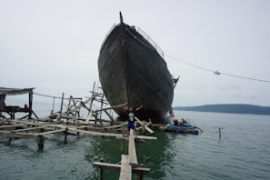 A large wooden ship is in the process of being built or repaired on a complex structure of wooden scaffolding over the water. A person is standing with arms outstretched on a narrow wooden plank leading to the ship. The scene is set against a calm body of water with a hill visible in the background.