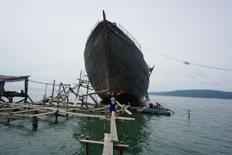A large wooden ship is in the process of being built or repaired on a complex structure of wooden scaffolding over the water. A person is standing with arms outstretched on a narrow wooden plank leading to the ship. The scene is set against a calm body of water with a hill visible in the background.