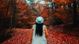 A person wearing a white fur vest, orange sleeves, and a light blue knit hat stands on a path covered with fallen red leaves, surrounded by trees with autumn foliage.