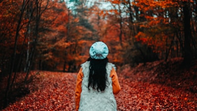 A person wearing a white fur vest, orange sleeves, and a light blue knit hat stands on a path covered with fallen red leaves, surrounded by trees with autumn foliage.