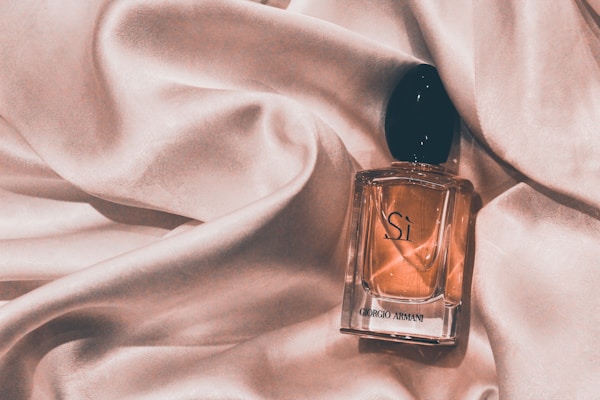 A perfume bottle labeled 'Sí Giorgio Armani' rests on a luxurious, light pink satin fabric that drapes softly around it. The bottle is clear with a black cap, containing a light amber-colored liquid.