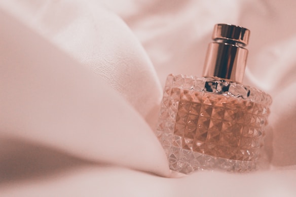 A clear, textured glass perfume bottle with a metallic cap is nestled in soft, white fabric. The fabric folds create a luxurious and elegant setting, complementing the sophisticated design of the bottle.