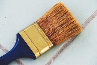 A large paintbrush with natural bristles and a metal ferrule can be seen lying on a tiled surface. The handle is dark blue, and the metal part is golden with the number 50 engraved on it. The background consists of light blue tiles with gray grout lines.