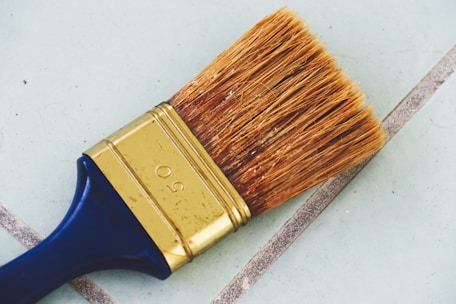 A large paintbrush with natural bristles and a metal ferrule can be seen lying on a tiled surface. The handle is dark blue, and the metal part is golden with the number 50 engraved on it. The background consists of light blue tiles with gray grout lines.