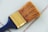 A large paintbrush with natural bristles and a metal ferrule can be seen lying on a tiled surface. The handle is dark blue, and the metal part is golden with the number 50 engraved on it. The background consists of light blue tiles with gray grout lines.