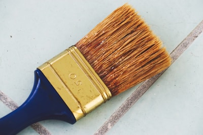 A large paintbrush with natural bristles and a metal ferrule can be seen lying on a tiled surface. The handle is dark blue, and the metal part is golden with the number 50 engraved on it. The background consists of light blue tiles with gray grout lines.