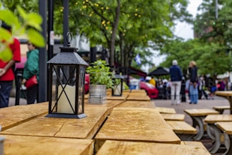 A black metal lantern with a candle inside is placed on a wooden outdoor table. In the background, people are gathered in a public space with trees and string lights visible. Green potted plants are also on the table, contributing to a cozy outdoor setting.