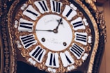 An antique clock with ornate gold detailing and Roman numerals on the clock face. The hands of the clock are elaborately designed, adding to the vintage aesthetic. The clock face has inscriptions in a cursive font.