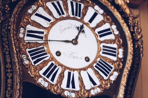 An antique clock with ornate gold detailing and Roman numerals on the clock face. The hands of the clock are elaborately designed, adding to the vintage aesthetic. The clock face has inscriptions in a cursive font.