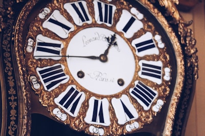 An antique clock with ornate gold detailing and Roman numerals on the clock face. The hands of the clock are elaborately designed, adding to the vintage aesthetic. The clock face has inscriptions in a cursive font.