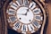An antique clock with ornate gold detailing and Roman numerals on the clock face. The hands of the clock are elaborately designed, adding to the vintage aesthetic. The clock face has inscriptions in a cursive font.