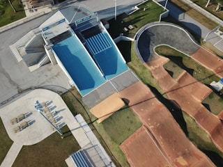 An aerial view of an outdoor sports facility featuring a blue skateboard ramp, a winding BMX track with dirt surfaces, and adjacent walking areas with seating and pathways. The surrounding area shows green grass and paved sections, with shadows indicating the presence of tall structures or lighting poles.