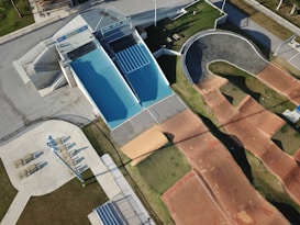 An aerial view of an outdoor sports facility featuring a blue skateboard ramp, a winding BMX track with dirt surfaces, and adjacent walking areas with seating and pathways. The surrounding area shows green grass and paved sections, with shadows indicating the presence of tall structures or lighting poles.