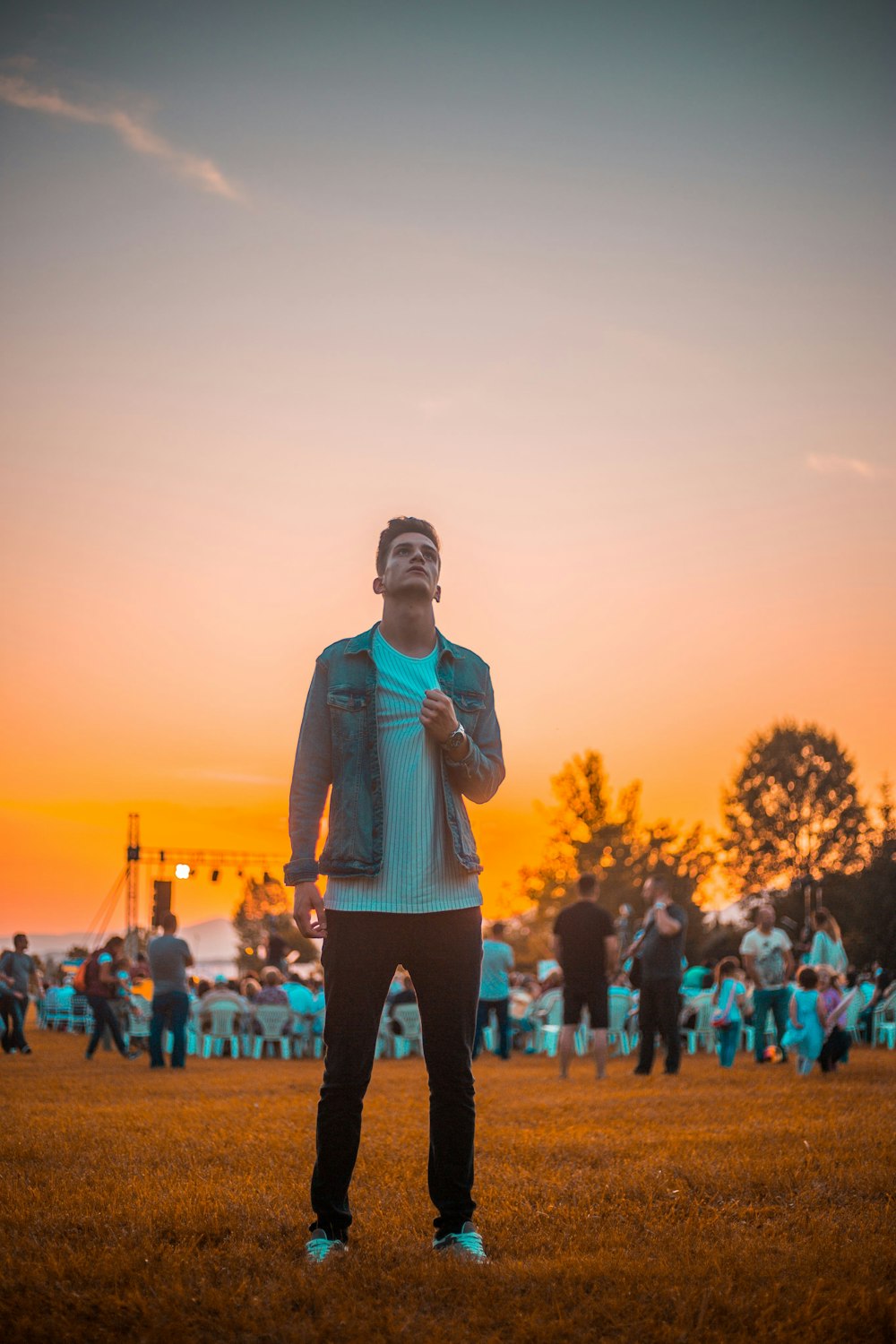 man wearing gray denim jacket standing on grass field while holding his white shirt and looking upward during golden hour