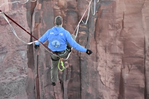 A person is daringly balancing on a tightrope across a deep canyon. The individual is wearing a blue hoodie and beige pants, with their back turned towards the camera, gazing at the steep rock formations of the canyon.