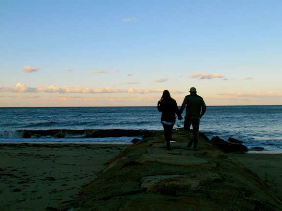 Couple holding hands walking on charcoal-hued rocks near the sea.