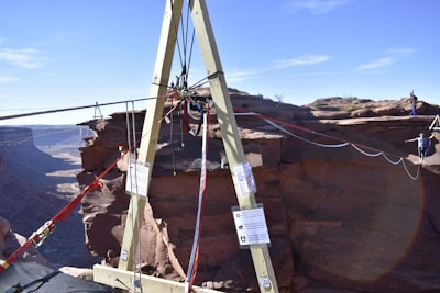 A wooden A-frame structure is set up on the edge of a canyon with ropes and rigging attached. In the background, a person is walking on a slackline that stretches across the canyon, highlighting a thrilling outdoor adventure scene.