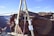 A wooden A-frame structure is set up on the edge of a canyon with ropes and rigging attached. In the background, a person is walking on a slackline that stretches across the canyon, highlighting a thrilling outdoor adventure scene.