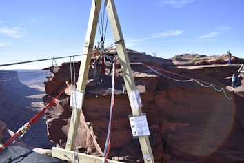 A wooden A-frame structure is set up on the edge of a canyon with ropes and rigging attached. In the background, a person is walking on a slackline that stretches across the canyon, highlighting a thrilling outdoor adventure scene.