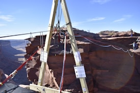 A wooden A-frame structure is set up on the edge of a canyon with ropes and rigging attached. In the background, a person is walking on a slackline that stretches across the canyon, highlighting a thrilling outdoor adventure scene.
