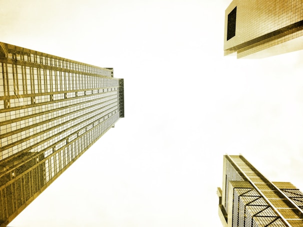 A perspective view between tall skyscrapers looking towards the sky. The buildings are reflective, with numerous windows arranged in a grid pattern. The architecture is modern and sleek, dominated by vertical and horizontal lines.