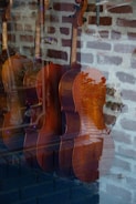 A reflection in a window displaying several string instruments, likely violins or violas, against a backdrop of a brick wall. The wood texture and rich brown color of the instruments are prominent, and there are reflections of trees and outdoor elements visible in the glass. The scene inside seems subdued and quietly elegant.
