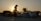 A person wearing a traditional Asian conical hat is silhouetted against the setting sun. They are standing next to a variety of packaged goods that are covered with plastic sheets. In the background, the sun is setting behind a palm tree and some rooftops.