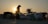 A person wearing a traditional Asian conical hat is silhouetted against the setting sun. They are standing next to a variety of packaged goods that are covered with plastic sheets. In the background, the sun is setting behind a palm tree and some rooftops.