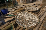 Circular woven trays are filled with fish laid out in a sun-drying arrangement. The trays are situated outdoors on a wooden platform, with various materials and containers scattered around. Buildings and fences can be seen in the background, indicating a village or small community setting.