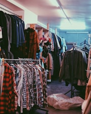 A cluttered room filled with clothes on hangers, ranging from plaid shirts to various jackets and coats. The setting appears to be a dimly lit second-hand or thrift store with narrow aisles between racks of clothing.