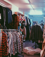 A cluttered room filled with clothes on hangers, ranging from plaid shirts to various jackets and coats. The setting appears to be a dimly lit second-hand or thrift store with narrow aisles between racks of clothing.