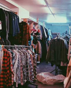 A cluttered room filled with clothes on hangers, ranging from plaid shirts to various jackets and coats. The setting appears to be a dimly lit second-hand or thrift store with narrow aisles between racks of clothing.