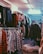 A cluttered room filled with clothes on hangers, ranging from plaid shirts to various jackets and coats. The setting appears to be a dimly lit second-hand or thrift store with narrow aisles between racks of clothing.