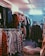 A cluttered room filled with clothes on hangers, ranging from plaid shirts to various jackets and coats. The setting appears to be a dimly lit second-hand or thrift store with narrow aisles between racks of clothing.