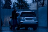 A person is washing a silver SUV at a car wash during the evening. The surroundings are dimly lit, with blue tones dominating the scene. The person is using a pressure washer to clean the vehicle. Trees and a corrugated metal fence are visible in the background.