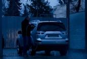 A person is washing a silver SUV at a car wash during the evening. The surroundings are dimly lit, with blue tones dominating the scene. The person is using a pressure washer to clean the vehicle. Trees and a corrugated metal fence are visible in the background.