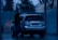 A person is washing a silver SUV at a car wash during the evening. The surroundings are dimly lit, with blue tones dominating the scene. The person is using a pressure washer to clean the vehicle. Trees and a corrugated metal fence are visible in the background.