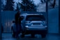 A person is washing a silver SUV at a car wash during the evening. The surroundings are dimly lit, with blue tones dominating the scene. The person is using a pressure washer to clean the vehicle. Trees and a corrugated metal fence are visible in the background.