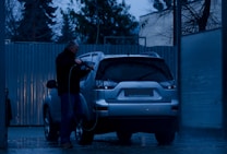 A person is washing a silver SUV at a car wash during the evening. The surroundings are dimly lit, with blue tones dominating the scene. The person is using a pressure washer to clean the vehicle. Trees and a corrugated metal fence are visible in the background.