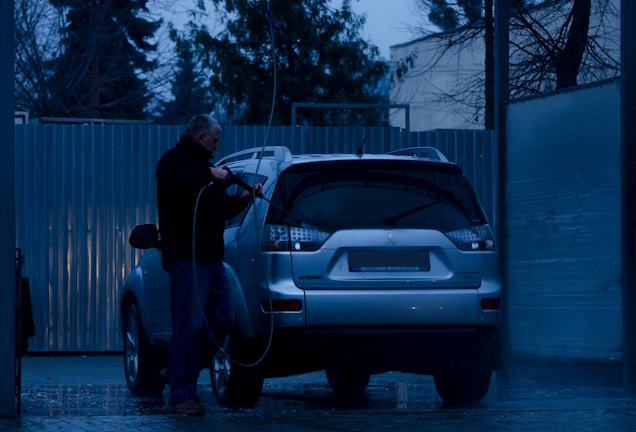 A person is washing a silver SUV at a car wash during the evening. The surroundings are dimly lit, with blue tones dominating the scene. The person is using a pressure washer to clean the vehicle. Trees and a corrugated metal fence are visible in the background.