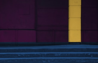 A minimalist wall with a striking vertical yellow stripe contrasting against a dark maroon background. The composition includes broad concrete steps leading up to the wall, adding an element of structure to the image.