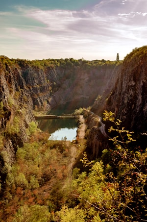 A picturesque landscape featuring a deep quarry with steep rocky walls surrounded by lush green trees. A narrow water body contrasts with the earthy tones of the cliffs, reflecting the sky above. The sun casts a warm, golden light over the scene, accentuating the textures of the rocks and foliage.
