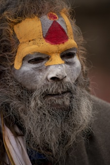 An elderly person with a long, grey beard and hair. Their face is elaborately painted with yellow, red, and white paint. The red and yellow paint forms a distinctive pattern on the forehead and around the eyes.
